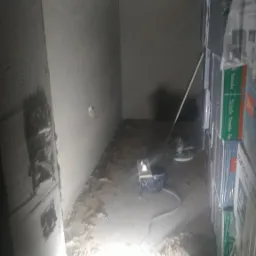 Wnętrze w trakcie prac tynkarskich: świeżo otynkowana ściana, wiadro, szlifierka do gładzi oraz zapas materiałów budowlanych w opakowaniach foliowych.