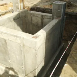 Surowy betonowy fundament w wykopie, widoczne ślady szalunku i metalowe elementy mocujące, ziemia wokół zabezpieczona taśmą ostrzegawczą.