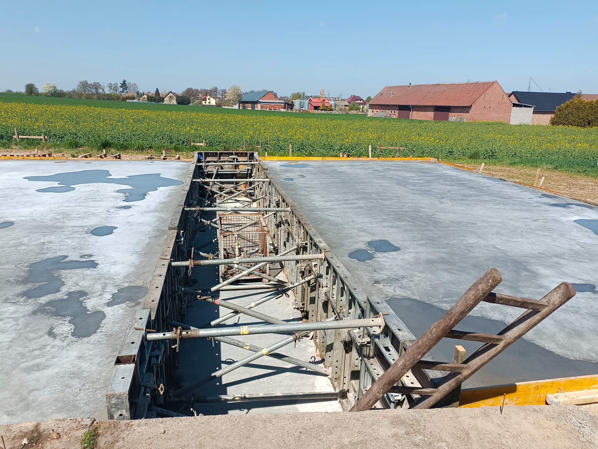 Wylany fundament z szalunkiem i drewnianą drabiną opartą o krawędź. W tle pola i zabudowania wiejskie w słoneczny dzień.