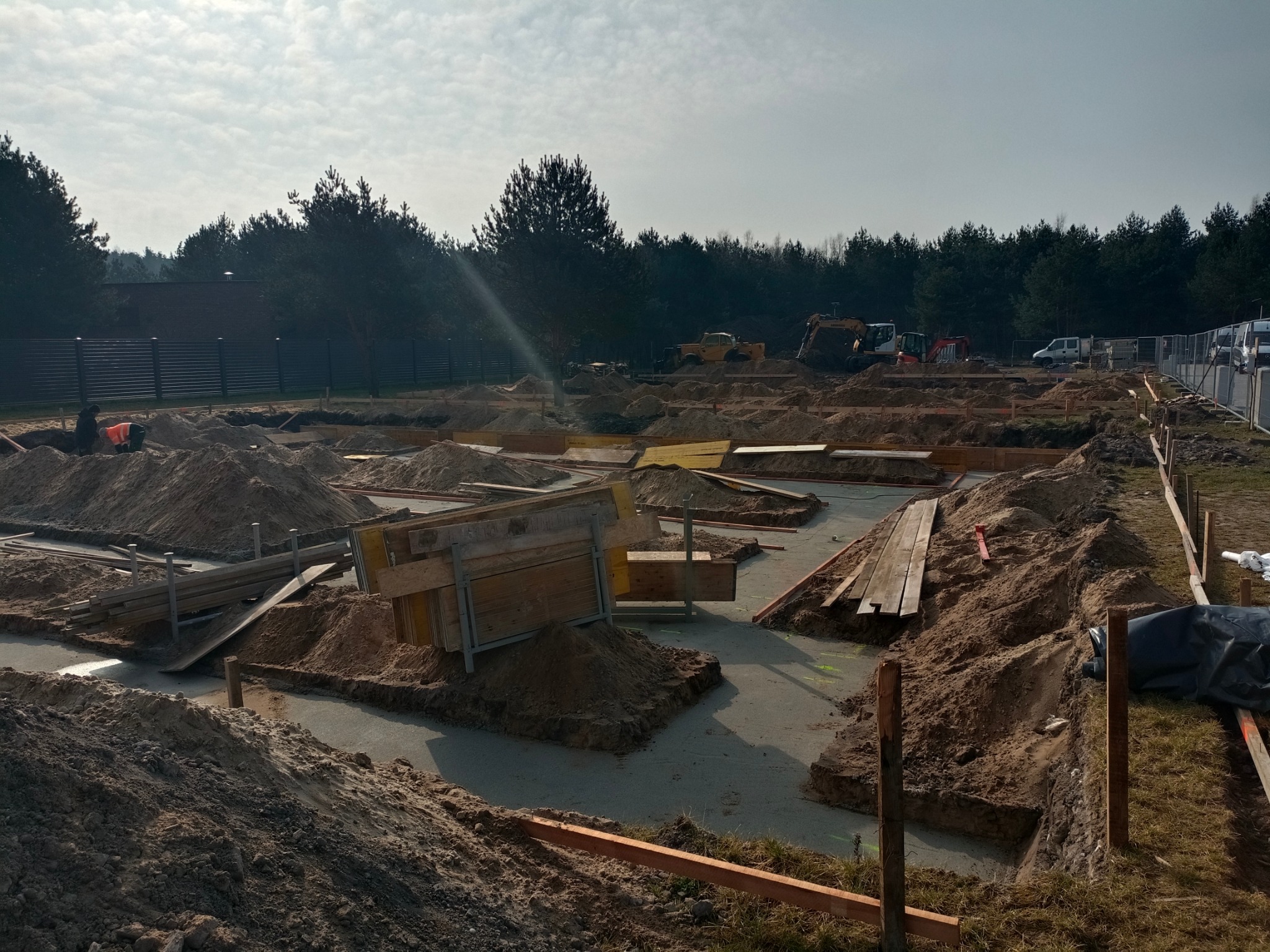 Wykonane wykopy pod fundamenty z częściowo wylanym betonem, widoczne szalunki i hałdy ziemi. W tle las i sprzęt budowlany na placu budowy.