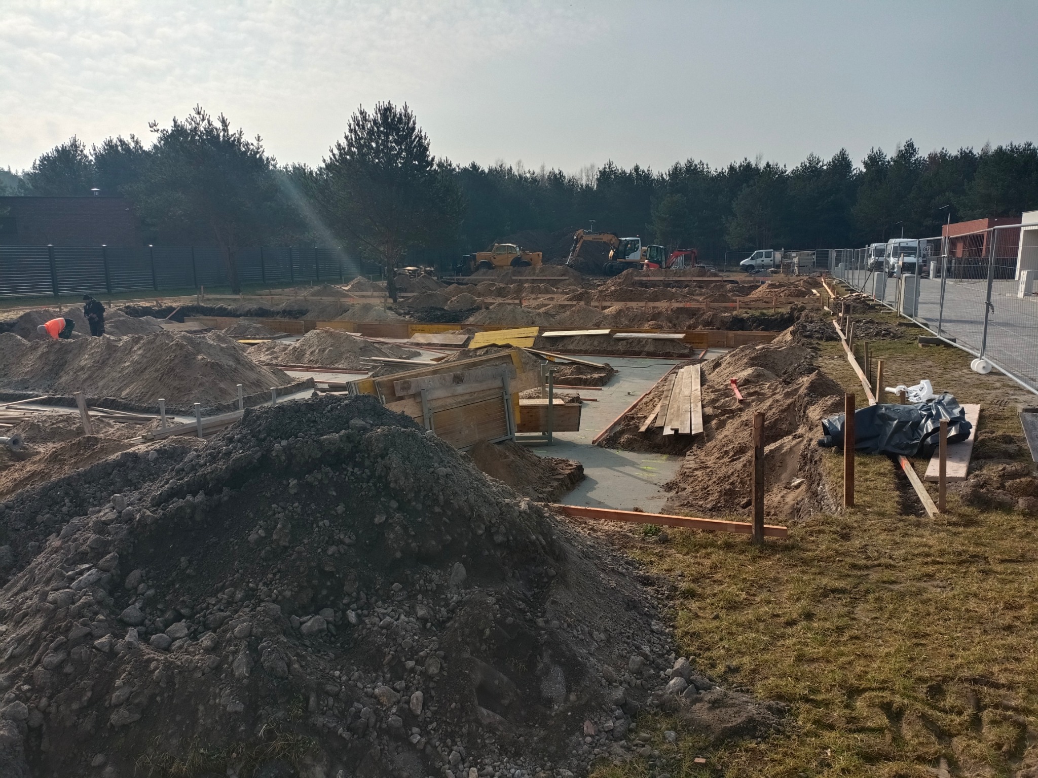 Wykop pod fundamenty domu w trakcie prac ziemnych. Widoczne szalunki, wykopane rowy, hałdy ziemi, sprzęt budowlany w tle i ogrodzenie terenu budowy.
