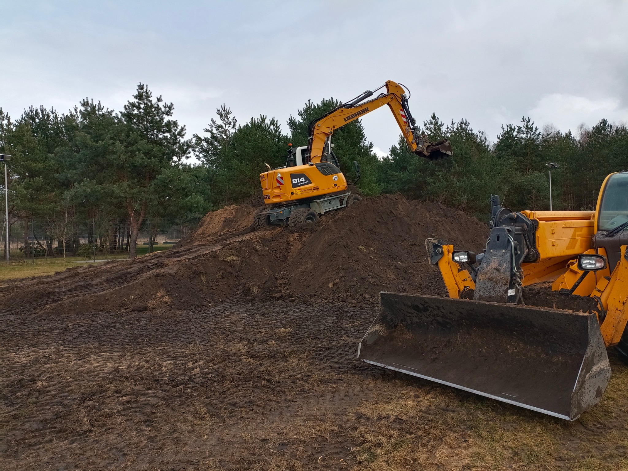 Żółta koparka Liebherr 914 Compact na usypanej ziemi, w tle las iglasty i pochmurne niebo. Widoczny fragment lemiesza spycharki.