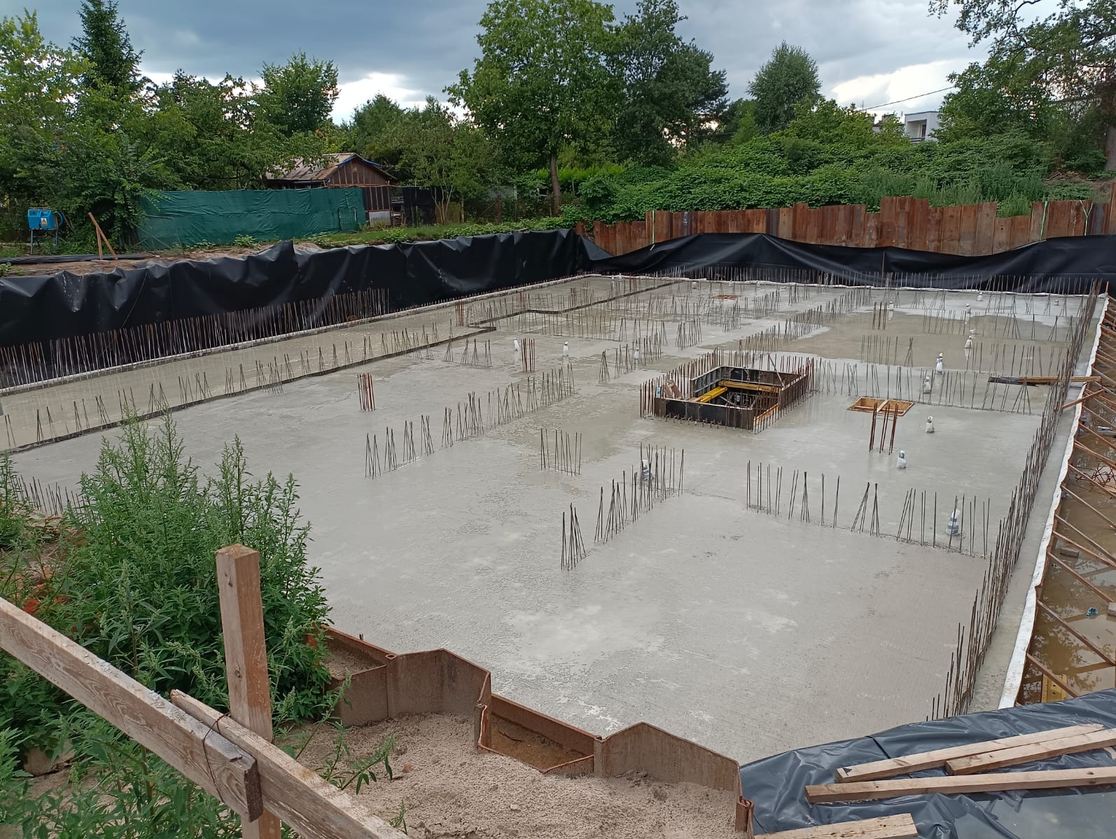 Zalany betonem fundament z wystającymi prętami zbrojeniowymi, zabezpieczony czarną folią. Widoczne szalunki i zarys przyszłych ścian. Tło stanowią drzewa i pochmurne niebo.