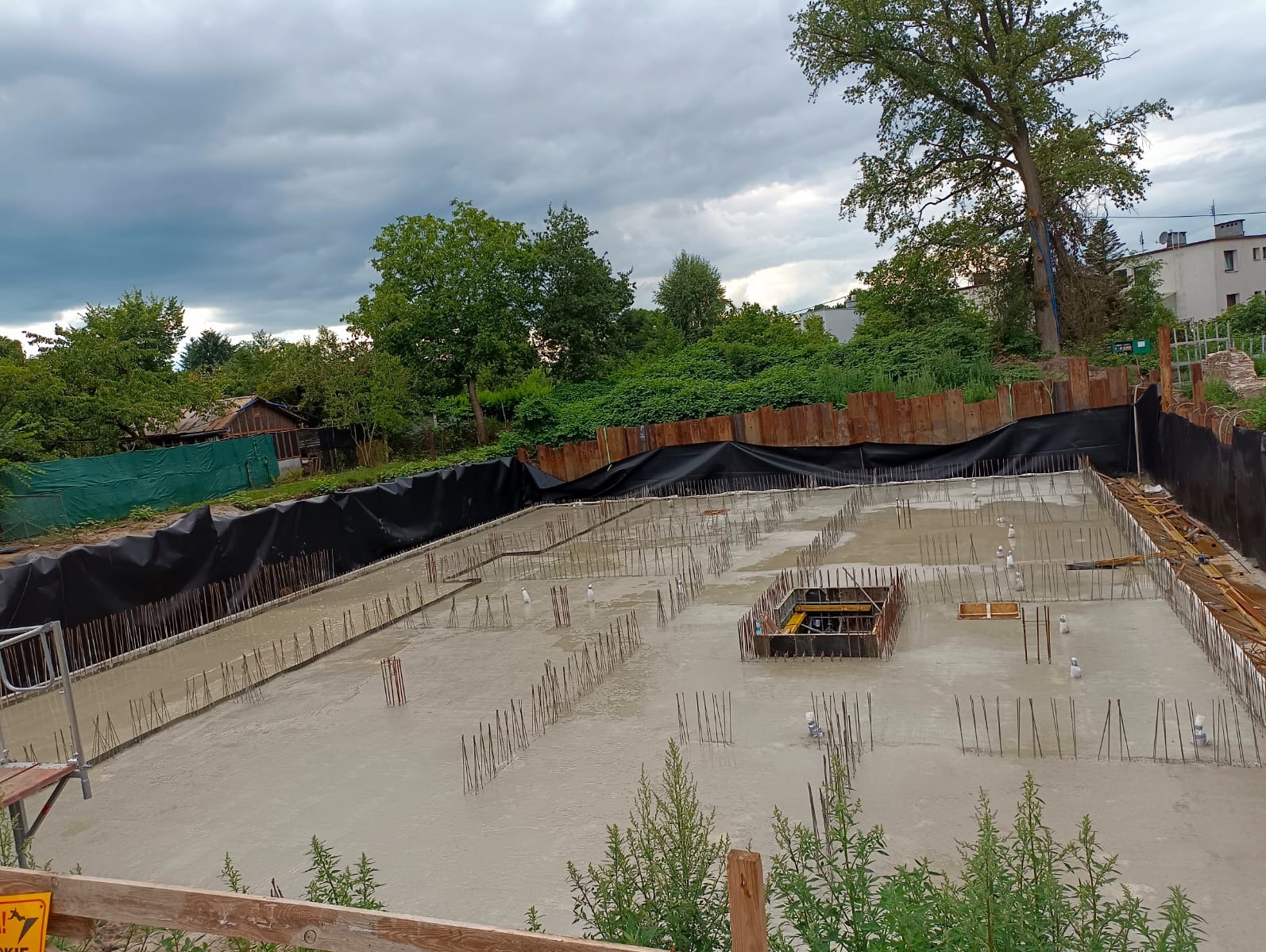 Zalany fundament z wystającymi prętami zbrojeniowymi, otoczony czarną folią i metalowymi panelami. W tle zieleń drzew i budynki pod pochmurnym niebem.
