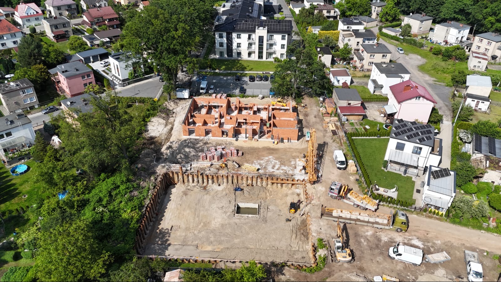 Zdjęcie z lotu ptaka przedstawia plac budowy z częściowo wzniesionymi ścianami z czerwonej cegły, zabezpieczonym wykopem i ciężkim sprzętem budowlanym. W tle zabudowa mieszkalna.