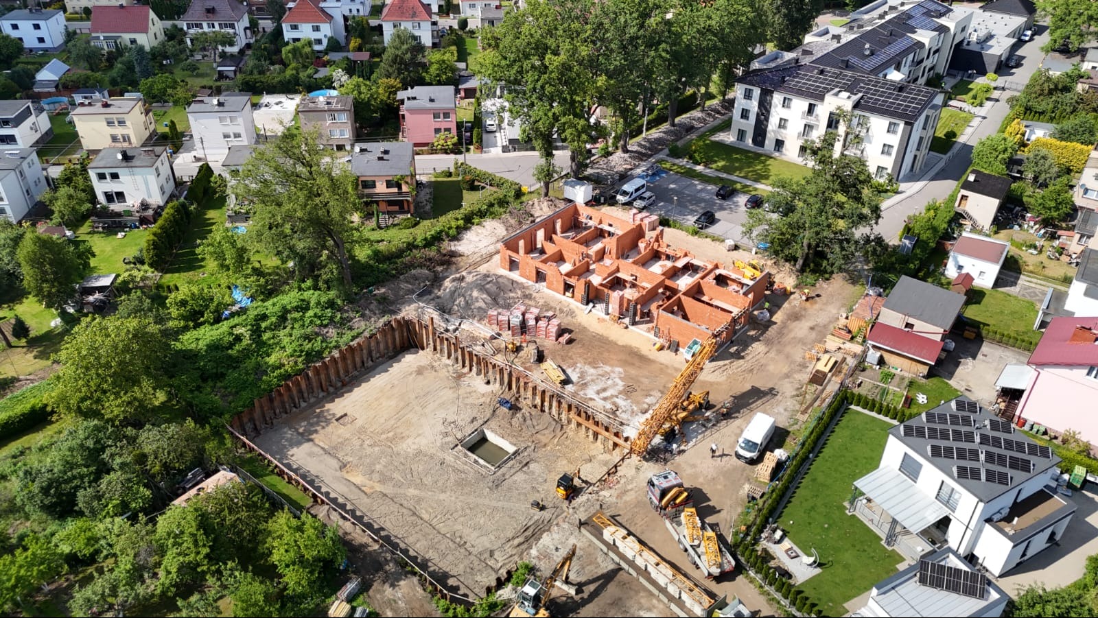 Widok z lotu ptaka na plac budowy z wykopem zabezpieczonym ściankami Larsena i wznoszoną konstrukcją domu z czerwonej cegły. Na placu budowy widoczne maszyny budowlane.