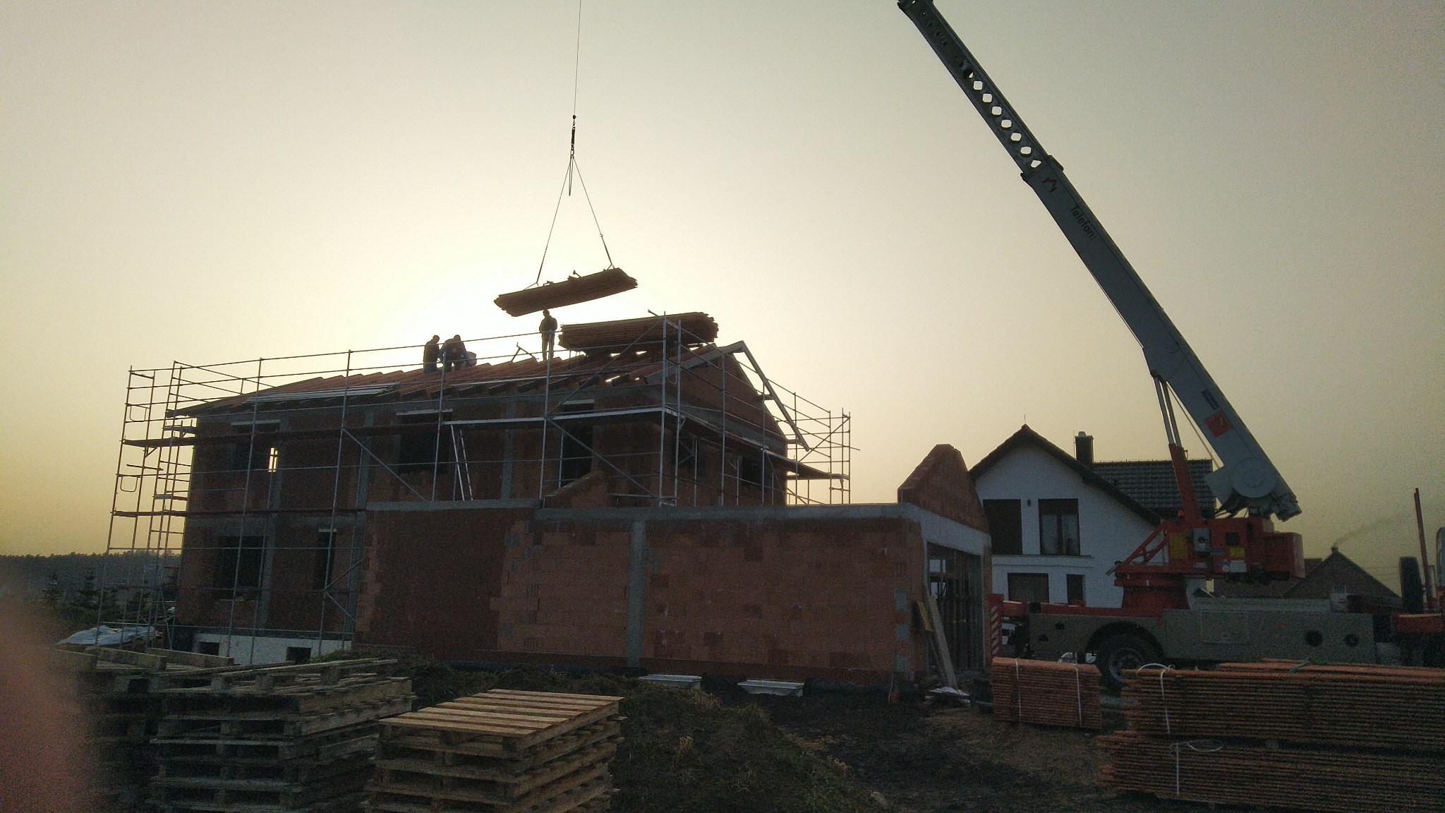 Budowa domu jednorodzinnego z czerwonej cegły: dźwig podnosi drewniane elementy więźby dachowej, robotnicy na rusztowaniu, w tle budynki mieszkalne.