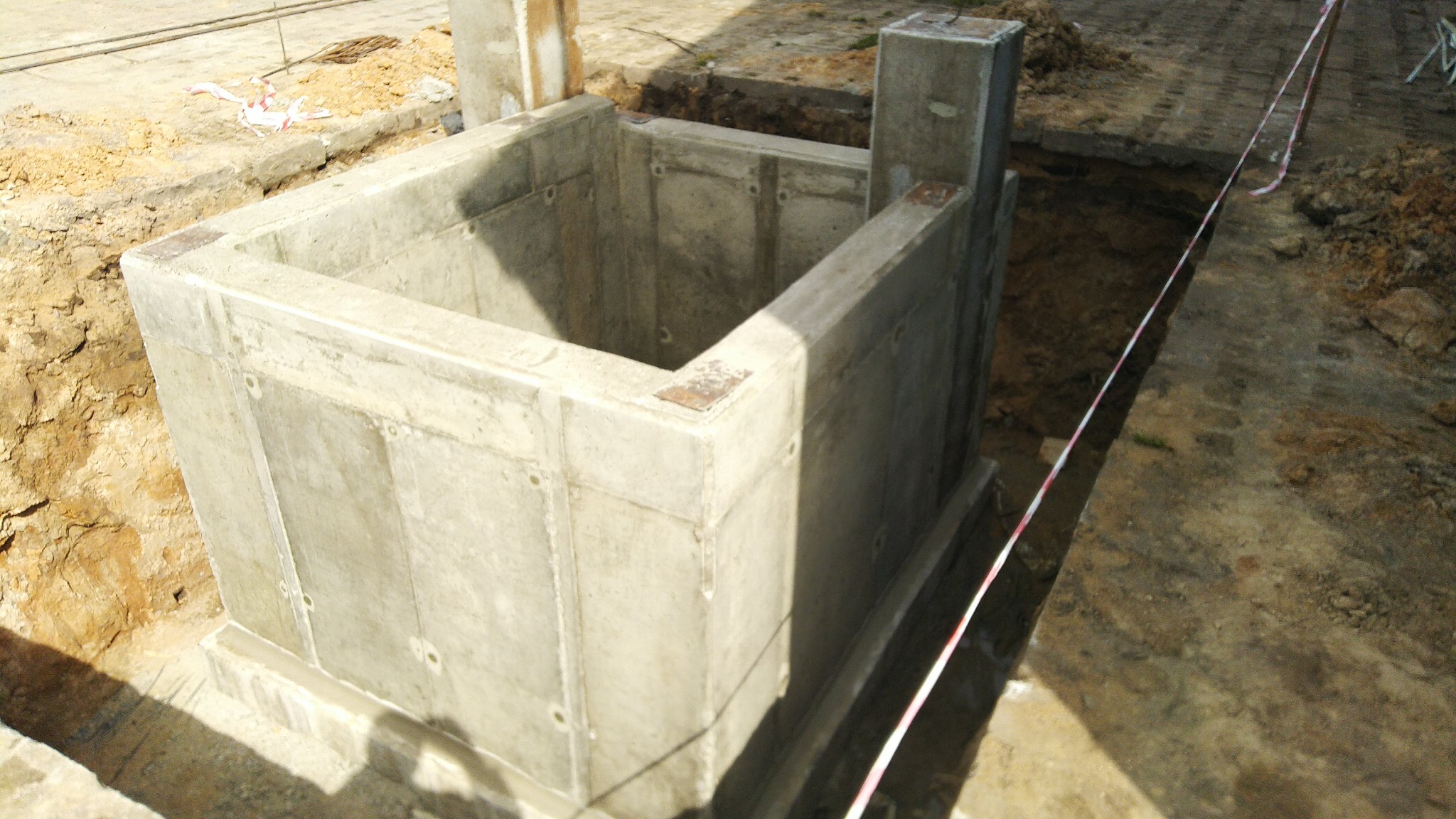 Surowy betonowy fundament w wykopie, widoczne ślady szalunku i metalowe elementy mocujące, ziemia wokół zabezpieczona taśmą ostrzegawczą.
