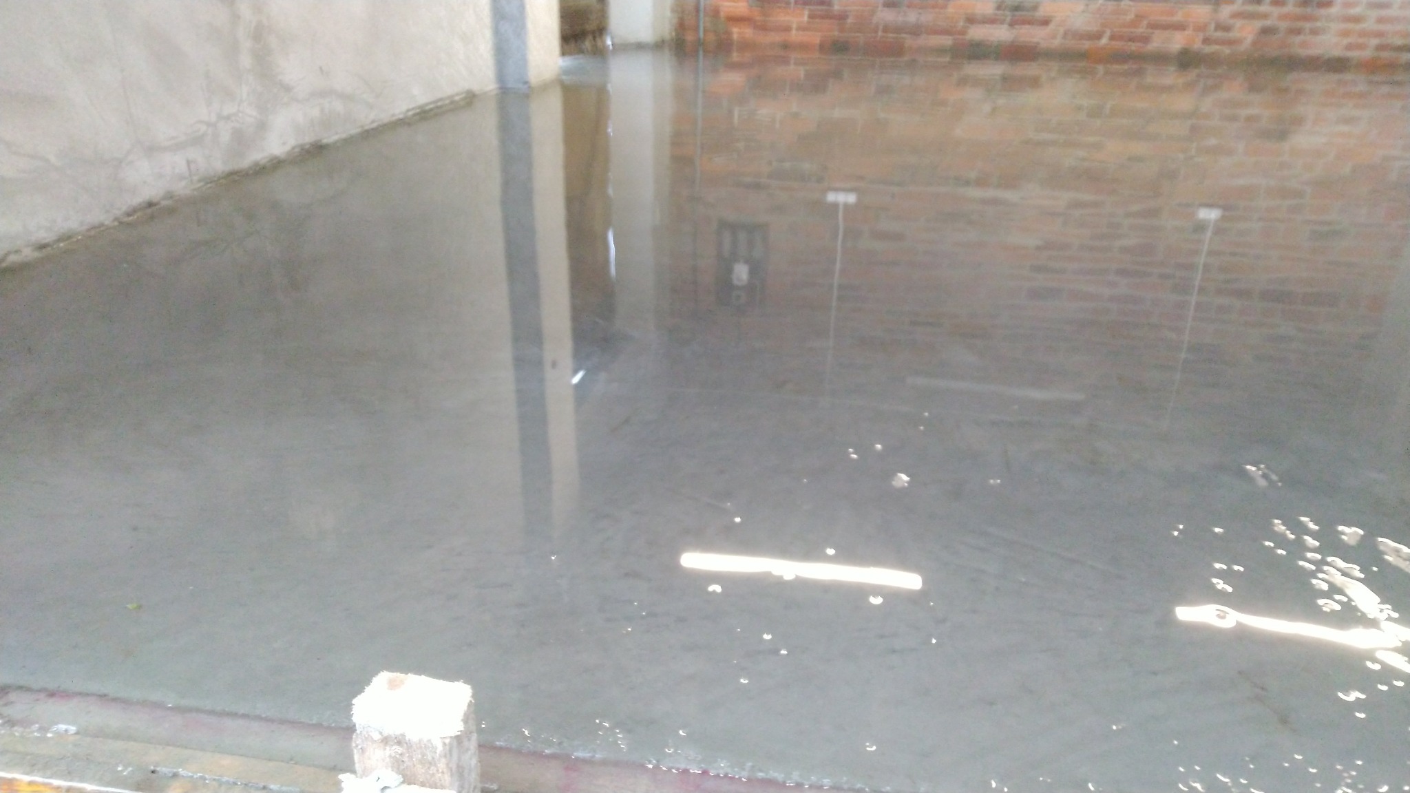 Świeżo wylana, mokra wylewka betonowa na podłodze, odbijająca światło, z widocznymi elementami szalunku i fragmentem ściany z czerwonej cegły w tle.