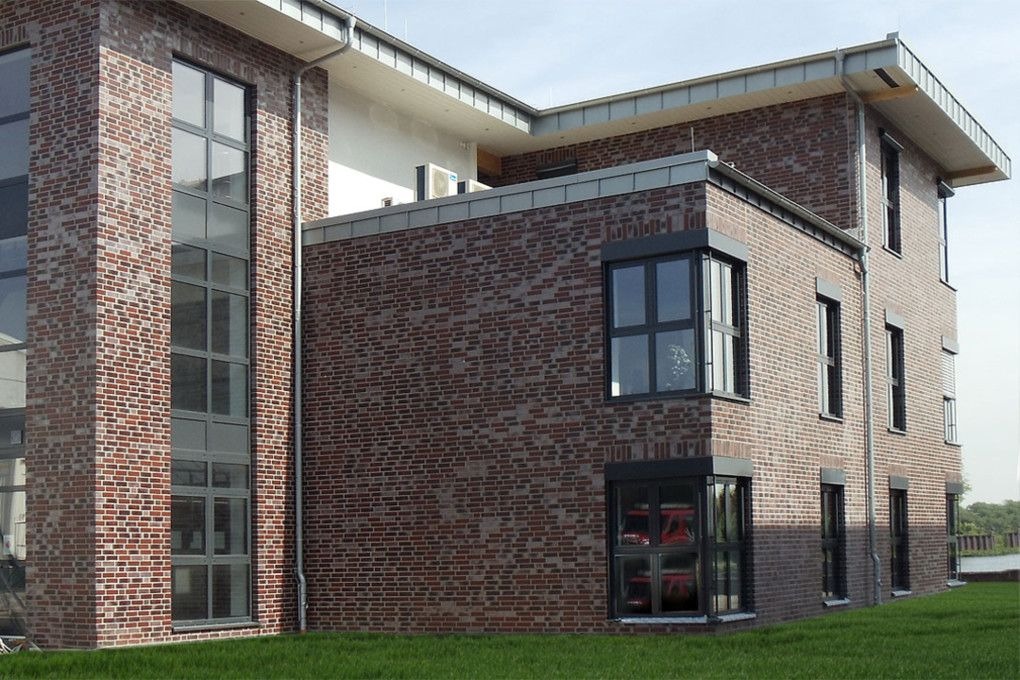 Nowoczesny, trzypiętrowy dom murowany z czerwonej cegły klinkierowej, z dużymi, grafitowymi oknami i metalowymi rynnami, otoczony zielonym trawnikiem.