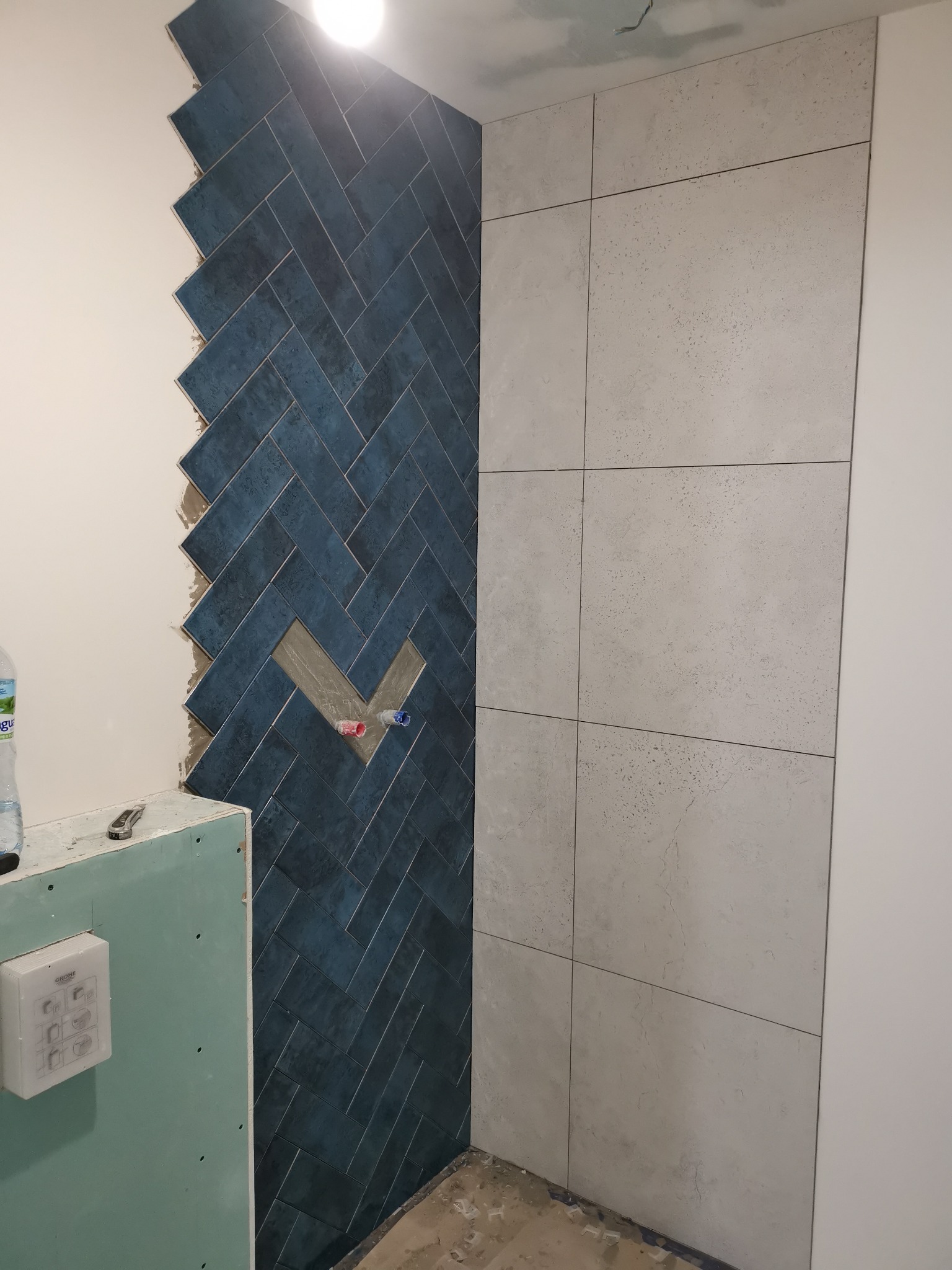 Montaż niebieskiej glazury w jodełkę na ścianie łazienki, obok ułożone duże, szare płytki, widoczne wystające rury i płyta karton-gips.