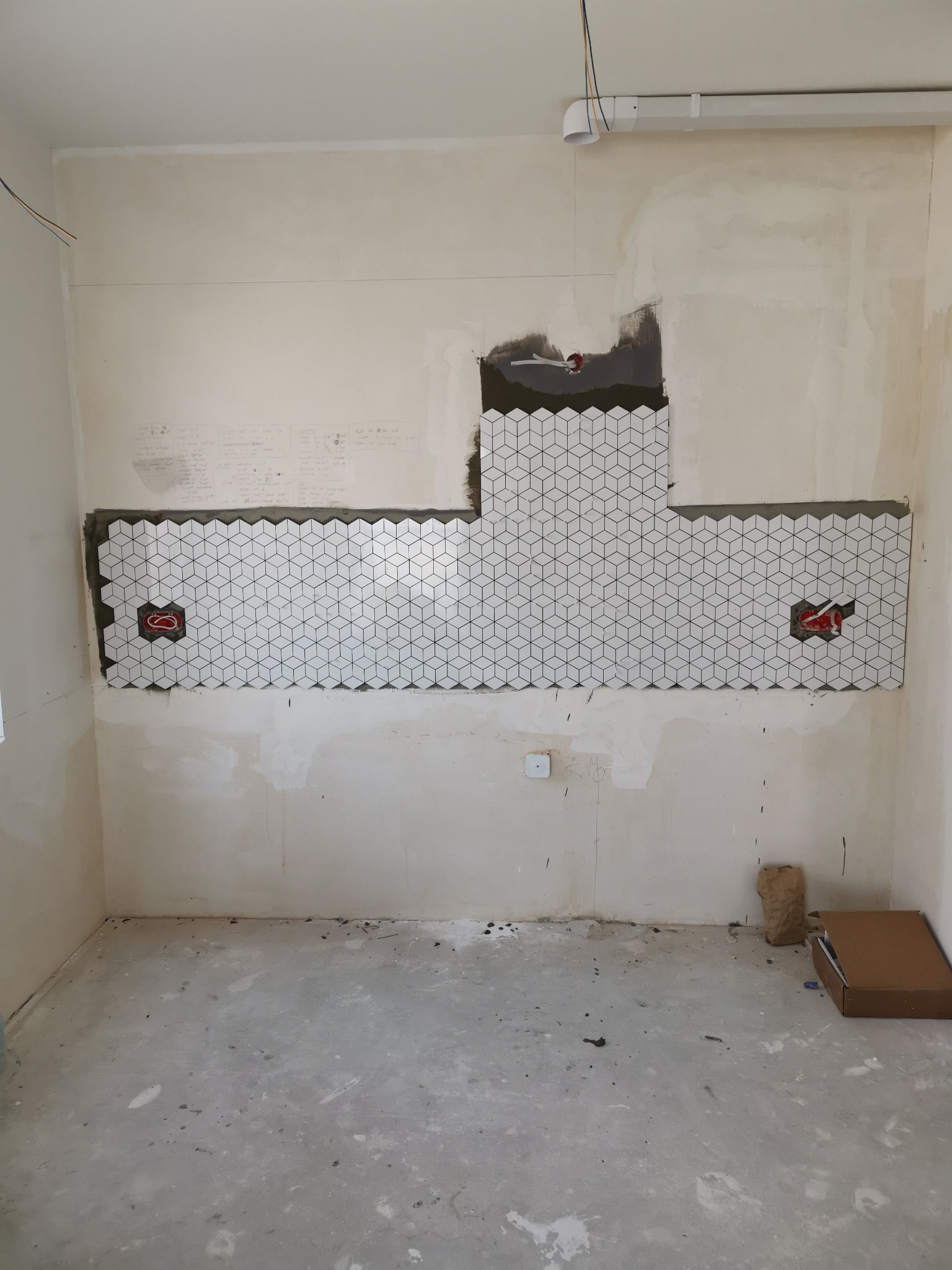 Ściana w trakcie układania mozaiki sześciennej, widoczne puszki elektryczne i niedokończone krawędzie, surowy stan podłogi i ścian.