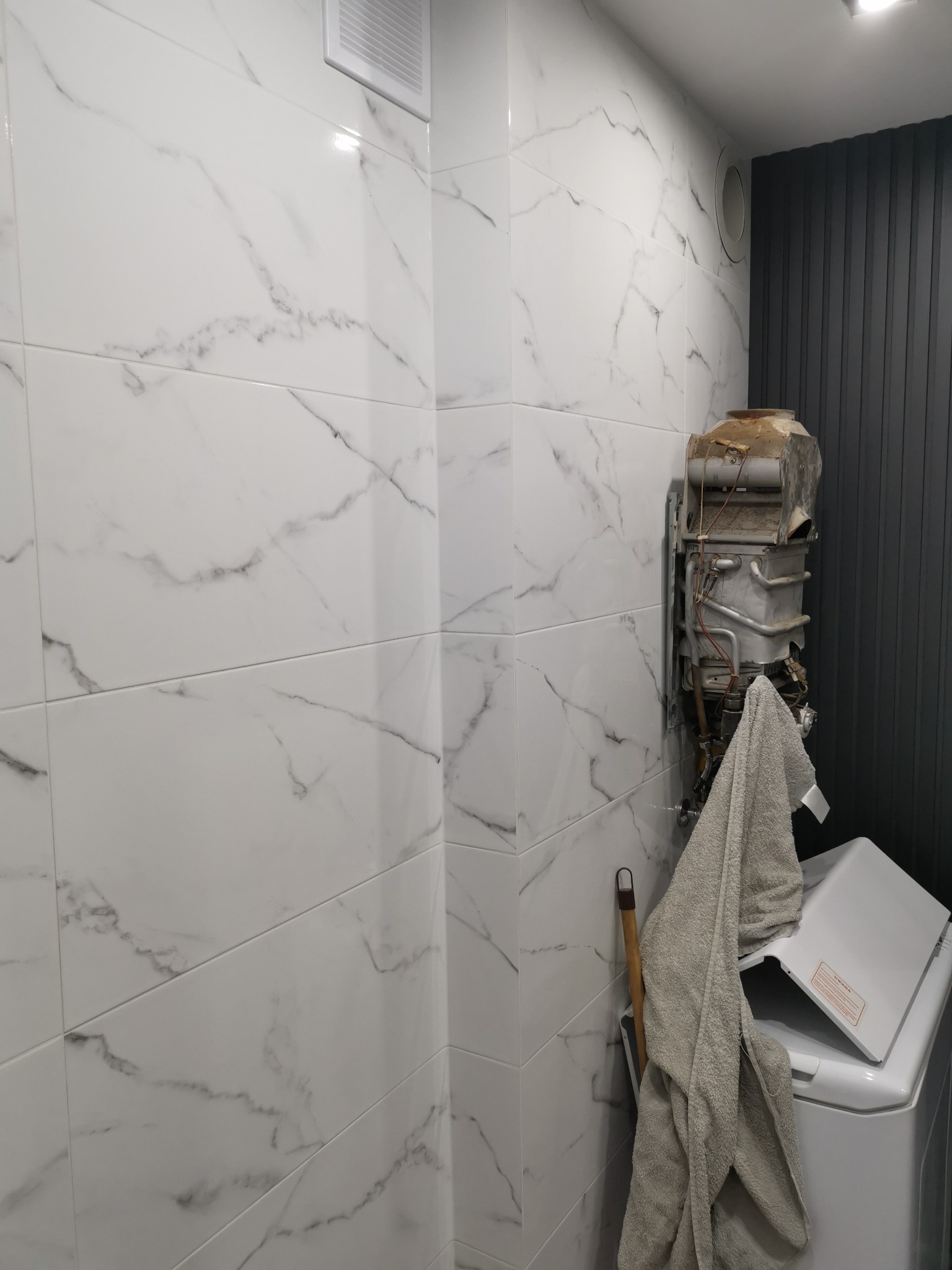 Łazienka w trakcie remontu: białe płytki imitujące marmur na ścianach, odsłonięty, stary i zardzewiały piec gazowy wiszący na ścianie, obok pralka z otwartą klapą i wiszący ręcznik.