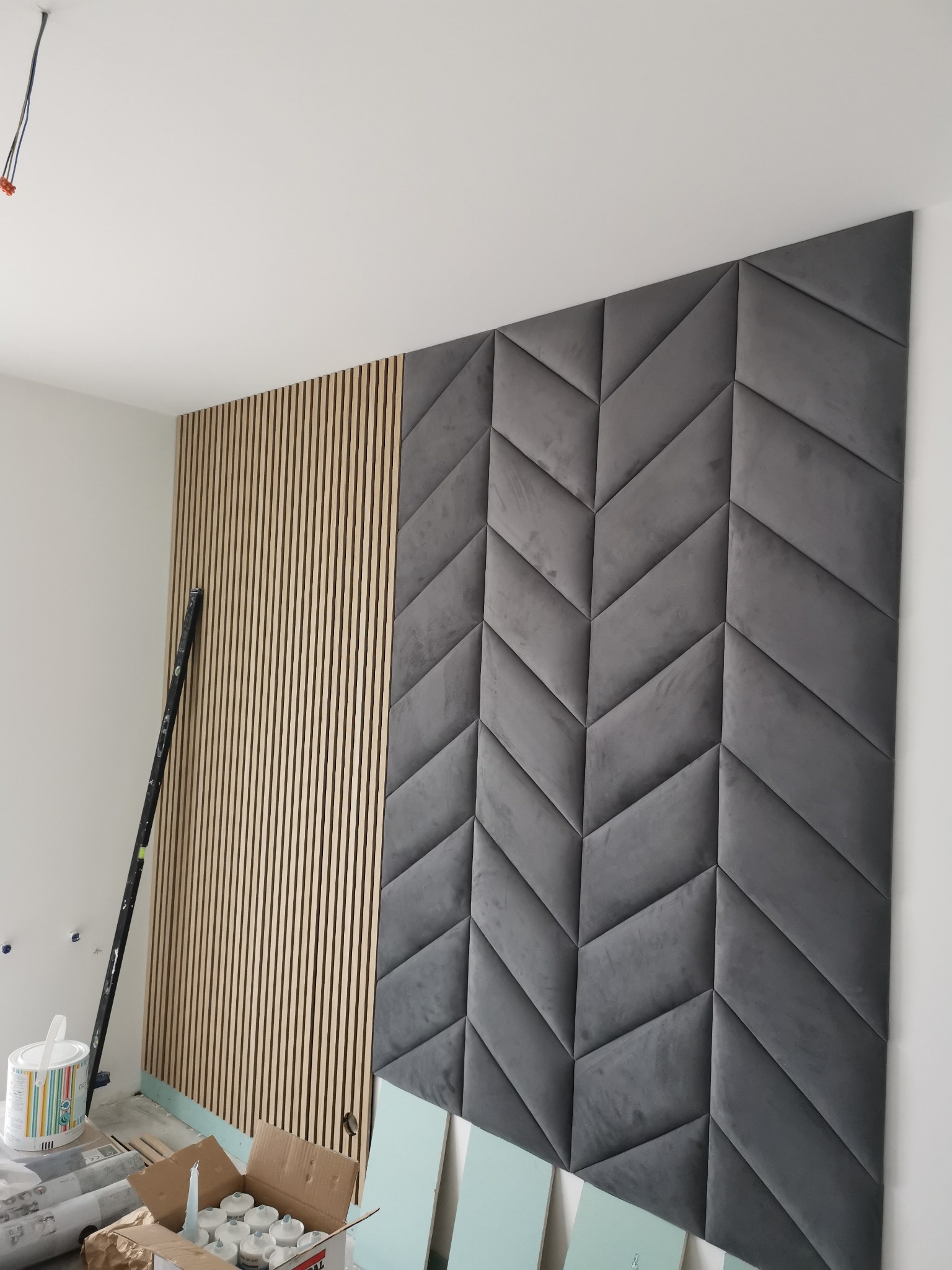 Montaż paneli dekoracyjnych: drewniane lamele i szare, welurowe panele w kształcie strzałek na ścianie, widoczne narzędzia i materiały budowlane.
