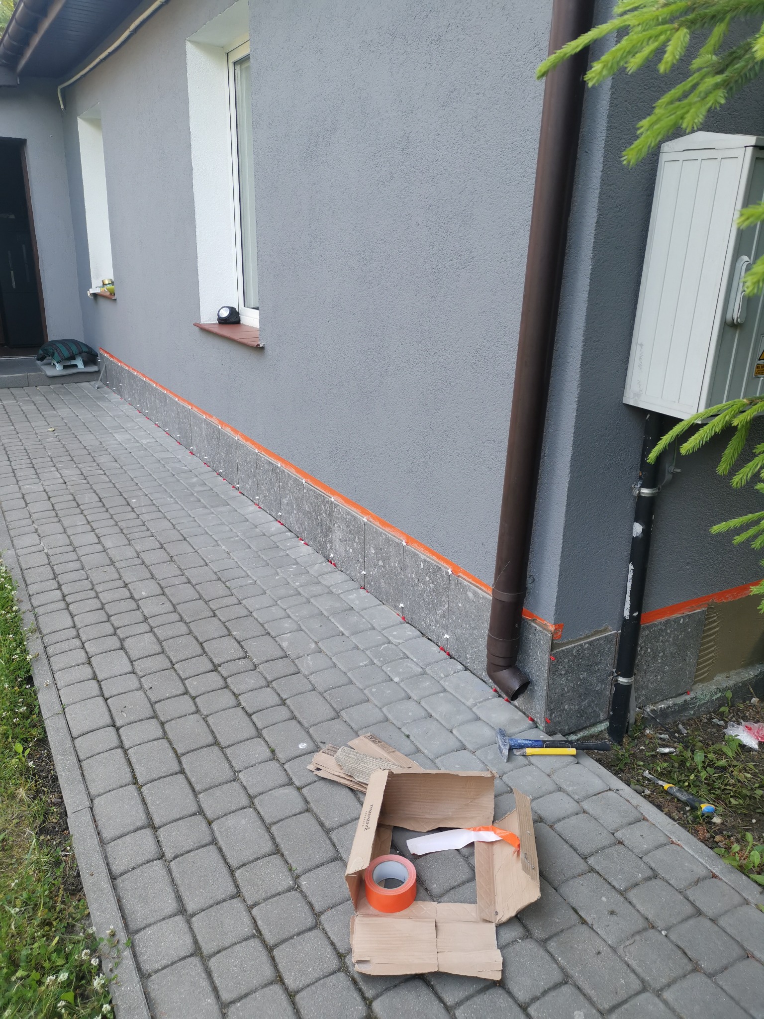 Przygotowanie do układania płytek elewacyjnych na cokole budynku z szarą elewacją, widoczne narzędzia, taśma malarska i krzyżyki dystansowe.