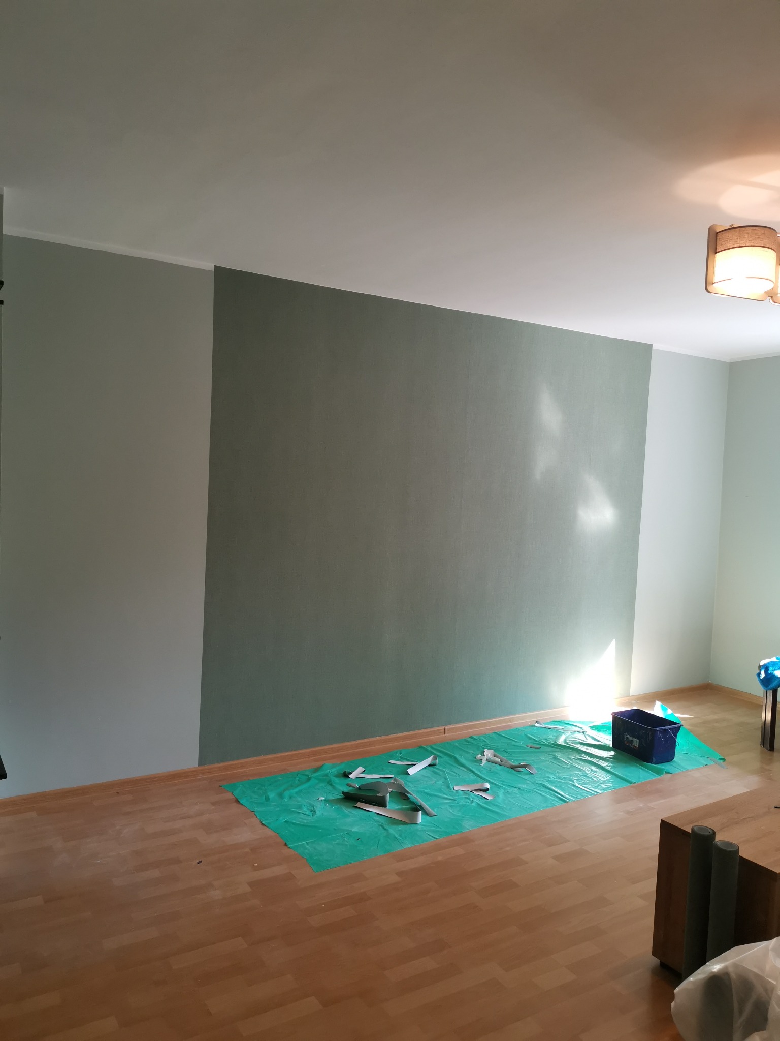 Pokój w trakcie malowania: ściana podzielona na dwa odcienie szarości, zabezpieczona podłoga folią malarską, widoczne resztki taśmy malarskiej i wiadro na podłodze.