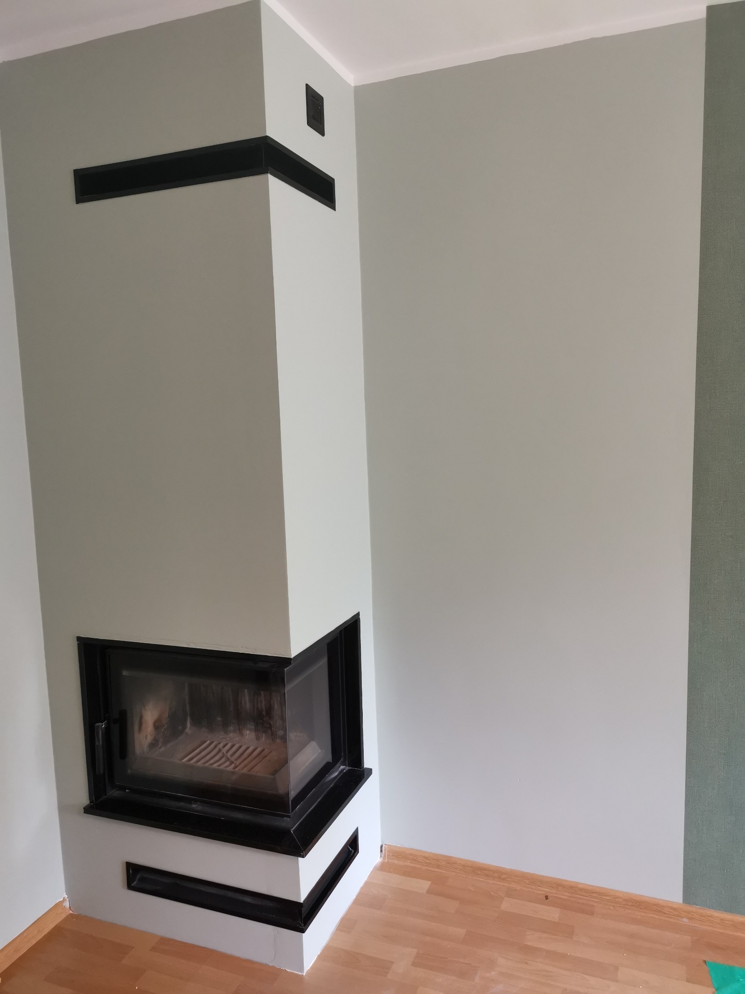 Nowoczesny narożny kominek z czarną obudową, wkomponowany w jasne ściany i drewnianą podłogę, z widocznymi elementami wentylacji w obudowie.