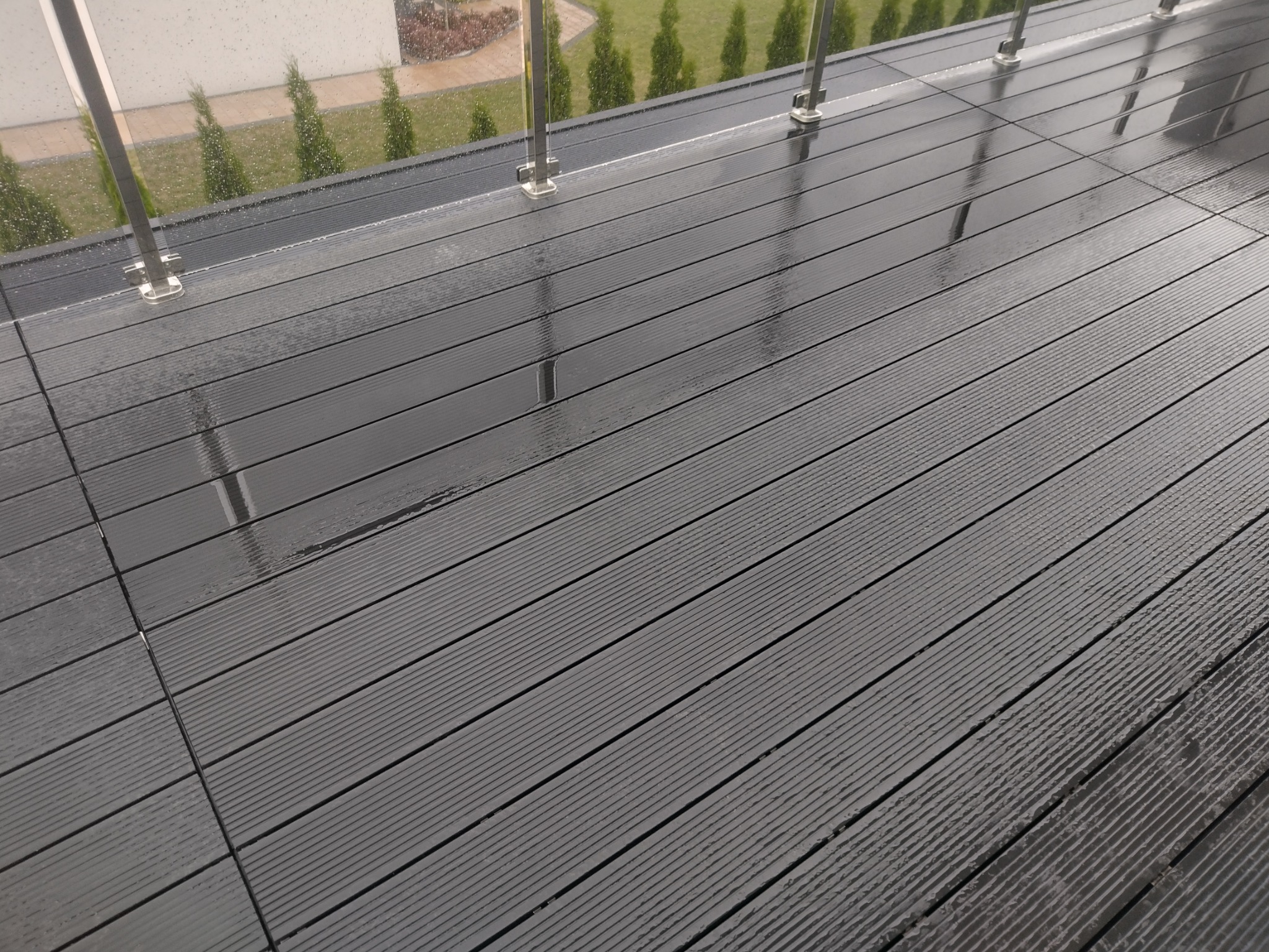 Deszcz na ciemnoszarym tarasie kompozytowym z balustradą ze szkła i stali nierdzewnej, widok na ogród z tujami.