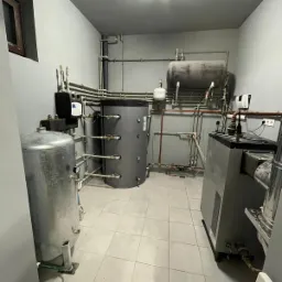 Kompleksowa kotłownia w szarej tonacji z zbiornikami, rurami miedzianymi i stalowymi, oraz kotłem grzewczym na tle kafelkowanej podłogi.