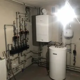 Kotłownia z piecem gazowym, zasobnikiem ciepłej wody i rozdzielaczem, widoczne liczne rury i zawory, na podłodze szare płytki, oświetlenie robocze.