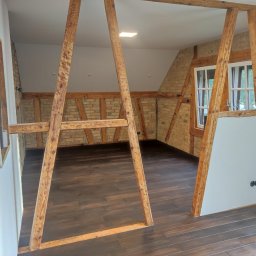 Sebastian warsz - Wnętrze poddasza z odsłoniętą konstrukcją szkieletową z belek, ceglanymi ścianami, nową podłogą z ciemnego drewna i przygotowanymi punktami elektrycznymi.