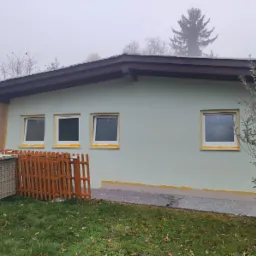 Ściana domu w trakcie tynkowania elewacji, zabezpieczone okna taśmą malarską, widoczne materiały budowlane i drewniane ogrodzenie.