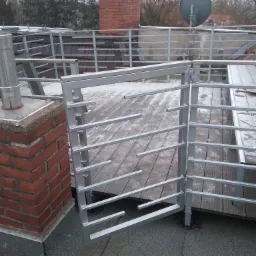 Szara metalowa balustrada na dachu z drewnianym tarasem, fragment komina z cegły i antena satelitarna w tle.