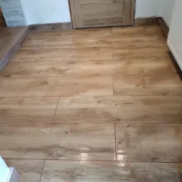 Układanie podłogi z płytek imitujących drewno w łazience, widoczna fuga w kolorze brązowym, drewniana listwa przypodłogowa i fragment grzejnika.