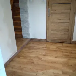 Ułożona podłoga z paneli imitujących drewno, widoczna drewniana ościeżnica drzwi z metalową klamką oraz fragment schodów z drewnianymi stopniami i ściana wyłożona szarymi płytkami imitującymi kamień.