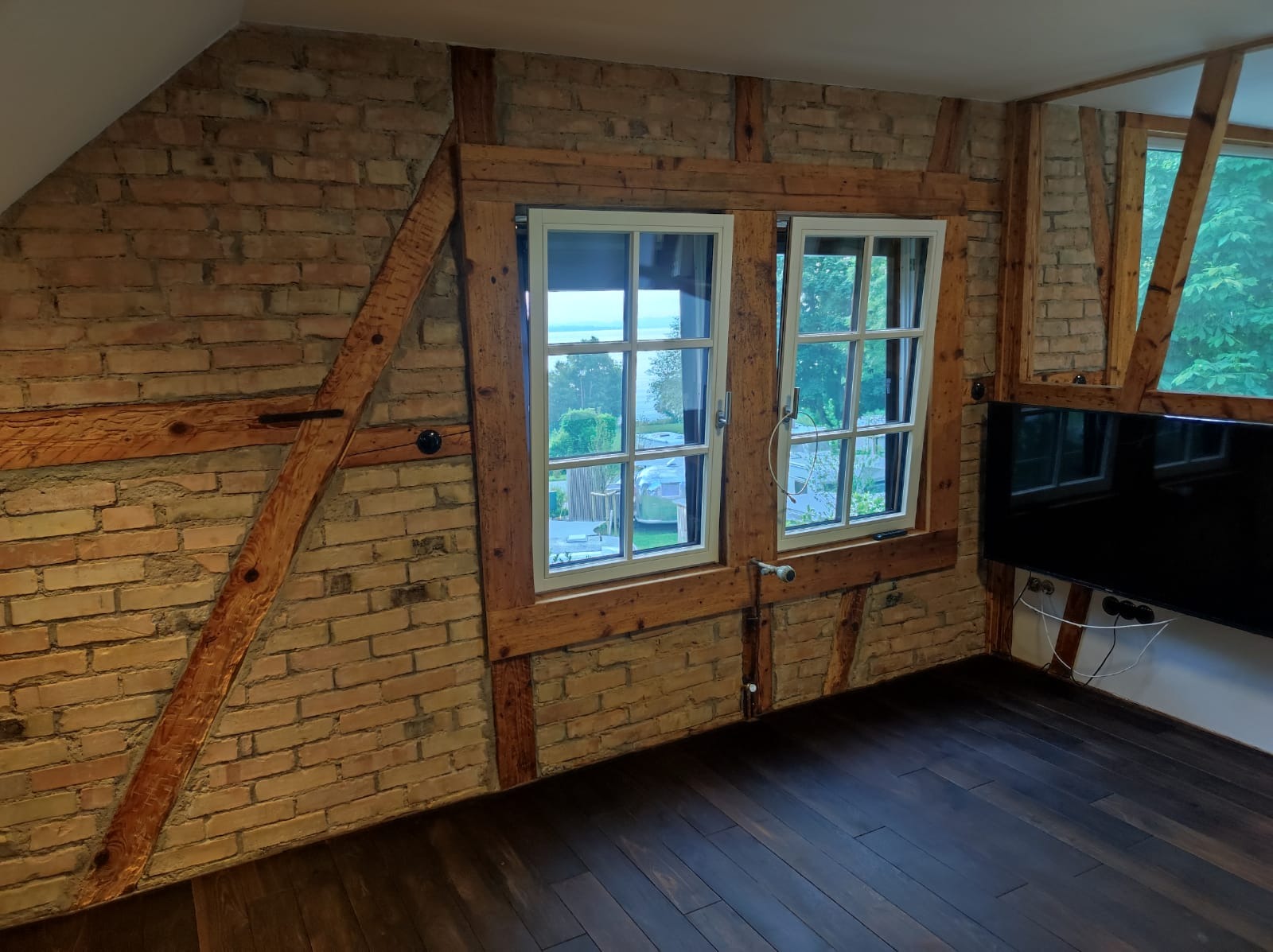 Poddasze z odsłoniętą cegłą, drewnianymi belkami konstrukcyjnymi i dwoma oknami z widokiem na zieleń, uzupełnione ciemną drewnianą podłogą i nowoczesnym telewizorem.
