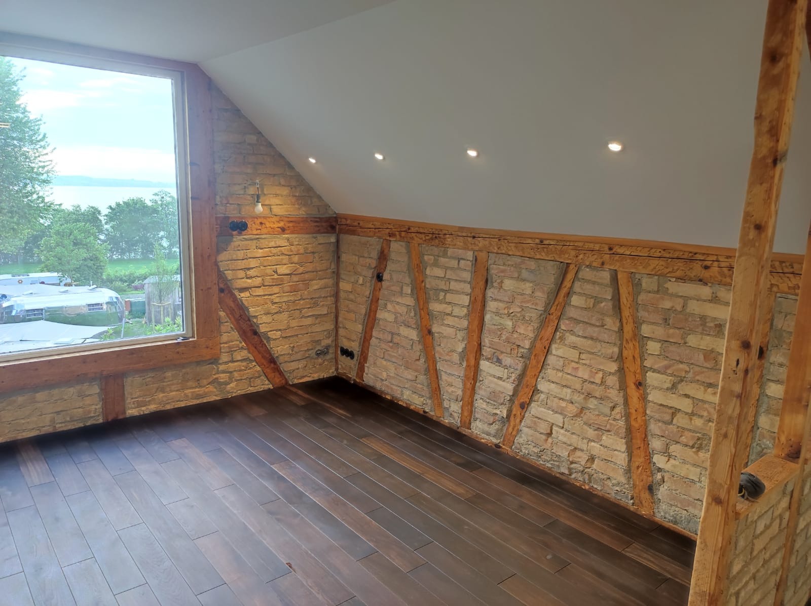 Poddasze z ceglaną ścianą, drewnianymi belkami konstrukcyjnymi i ciemną podłogą z desek. Widok na jezioro i zieleń za oknem.