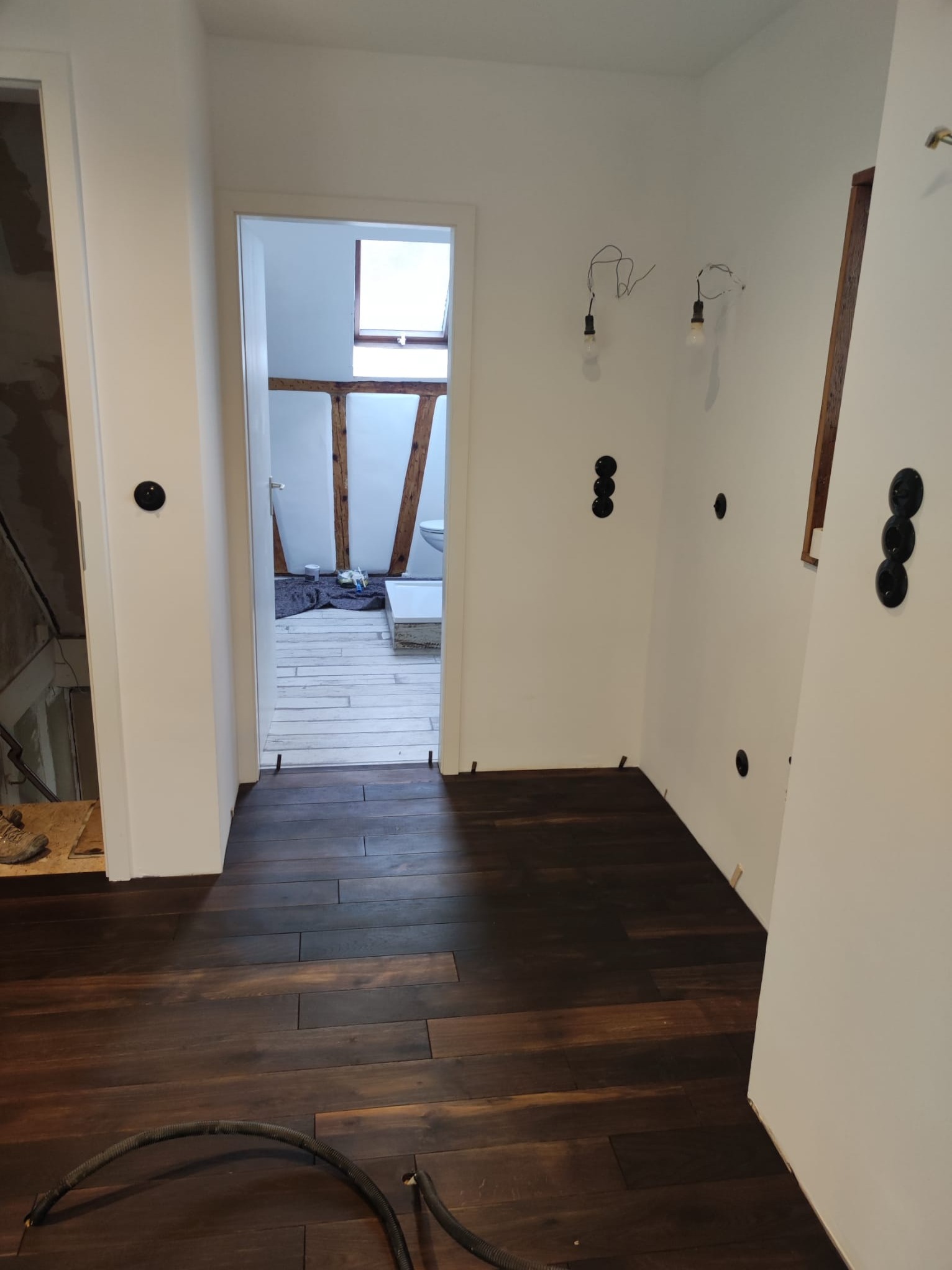 Wnętrze w trakcie prac wykończeniowych: ciemna drewniana podłoga, białe ściany z widocznymi puszkami elektrycznymi i wystającymi przewodami, w tle widoczna łazienka z drewnianymi elementami...