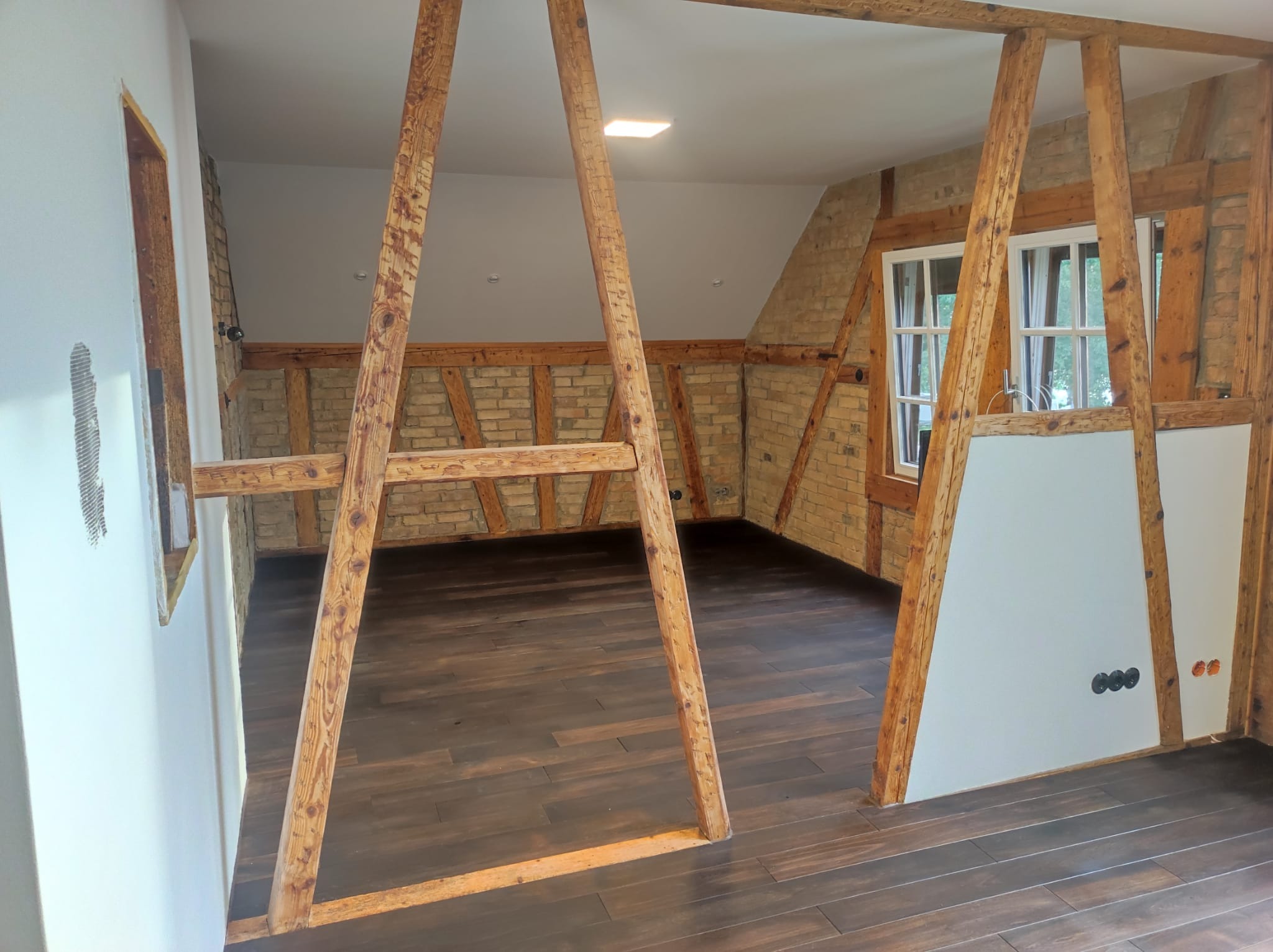 Wnętrze poddasza z odsłoniętą konstrukcją szkieletową z belek, ceglanymi ścianami, nową podłogą z ciemnego drewna i przygotowanymi punktami elektrycznymi.