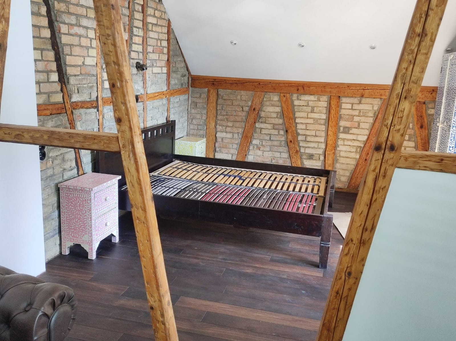 Pokój na poddaszu z odsłoniętą cegłą i drewnianymi belkami, z ciemną drewnianą ramą łóżka bez materaca i dekoracyjną szafką nocną w stylu boho.