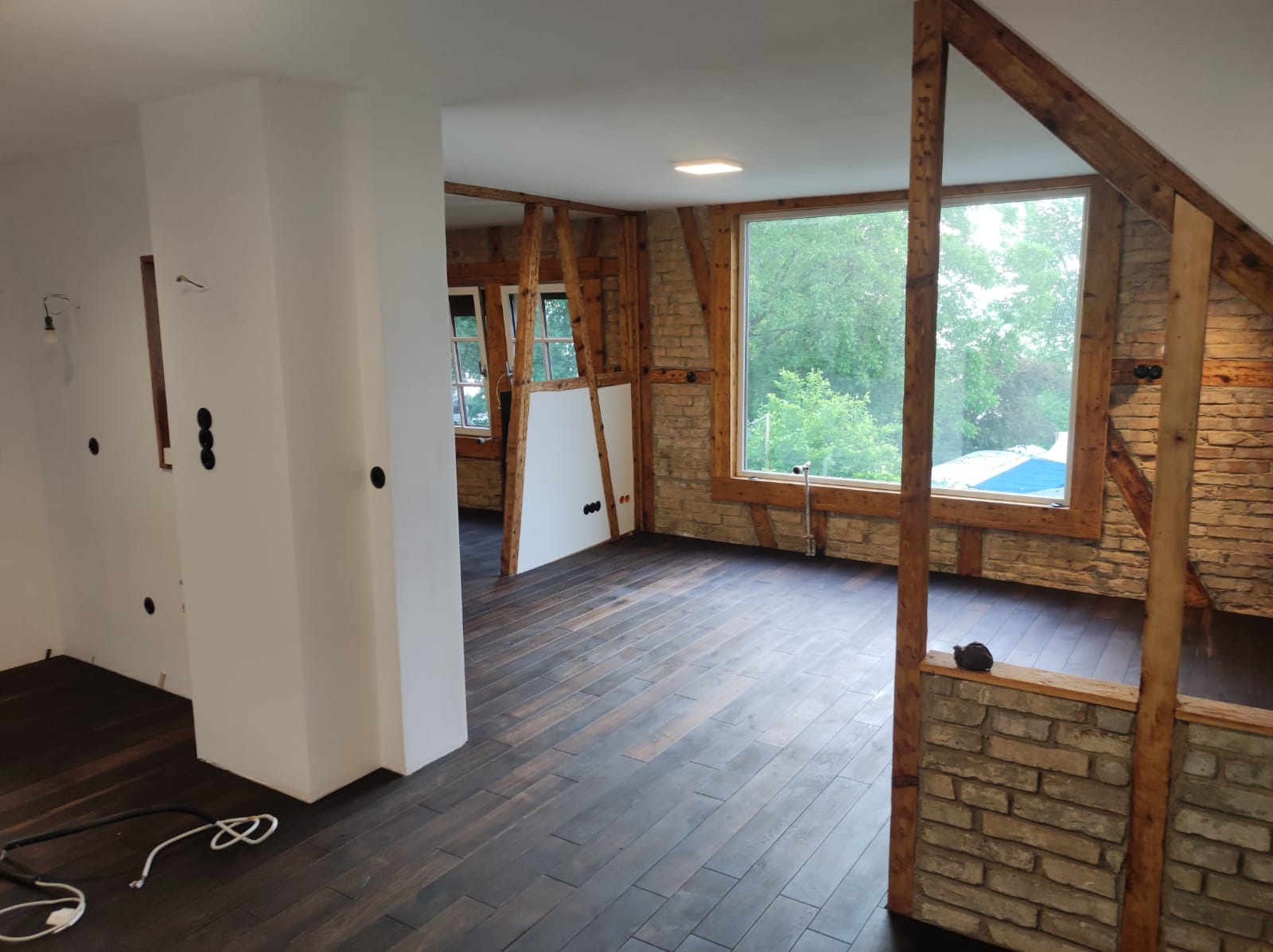 Wnętrze poddasza w trakcie remontu: białe ściany z czarnymi gniazdkami, drewniana konstrukcja nośna, okno z widokiem na zieleń, ciemna drewniana podłoga ułożona w deski.