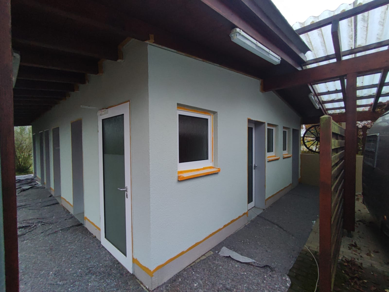 Budynek z zewnątrz, świeżo otynkowany na jasnoszaro, z pomarańczową taśmą malarską zabezpieczającą okna i drzwi, widoczne zadaszenie z drewnianych belek.