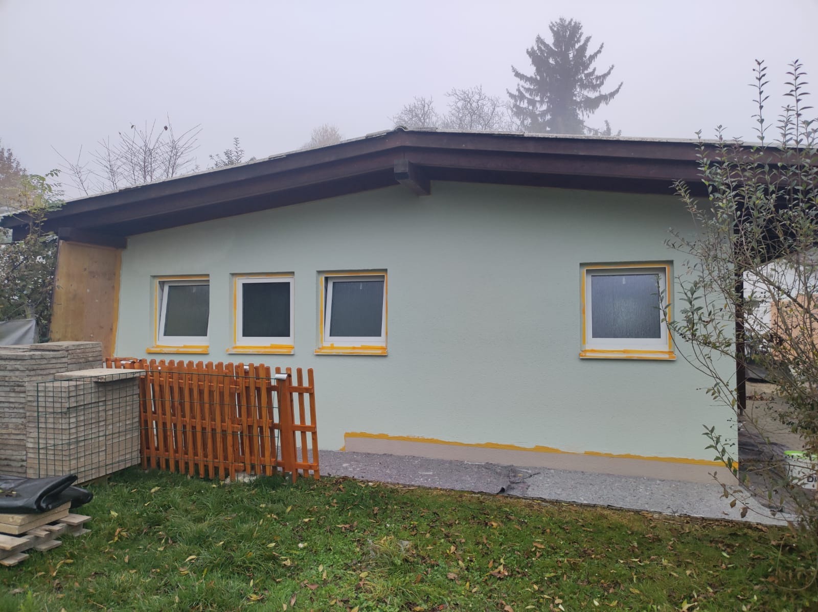 Ściana domu w trakcie tynkowania elewacji, zabezpieczone okna taśmą malarską, widoczne materiały budowlane i drewniane ogrodzenie.