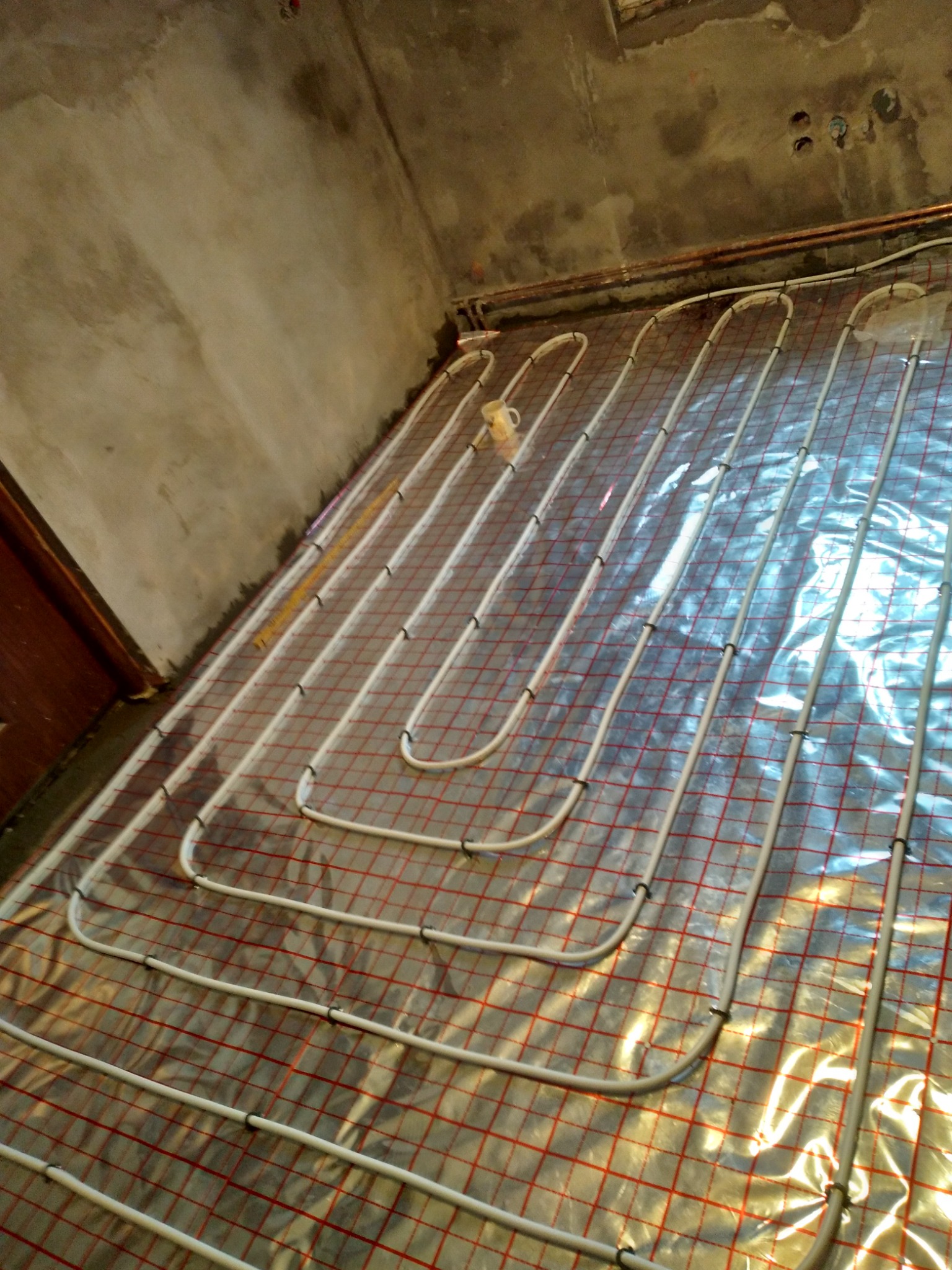 Instalacja ogrzewania podłogowego z rur na folii z czerwoną siatką, widoczne fragmenty surowych ścian i instalacji elektrycznej oraz kubek na podłodze.