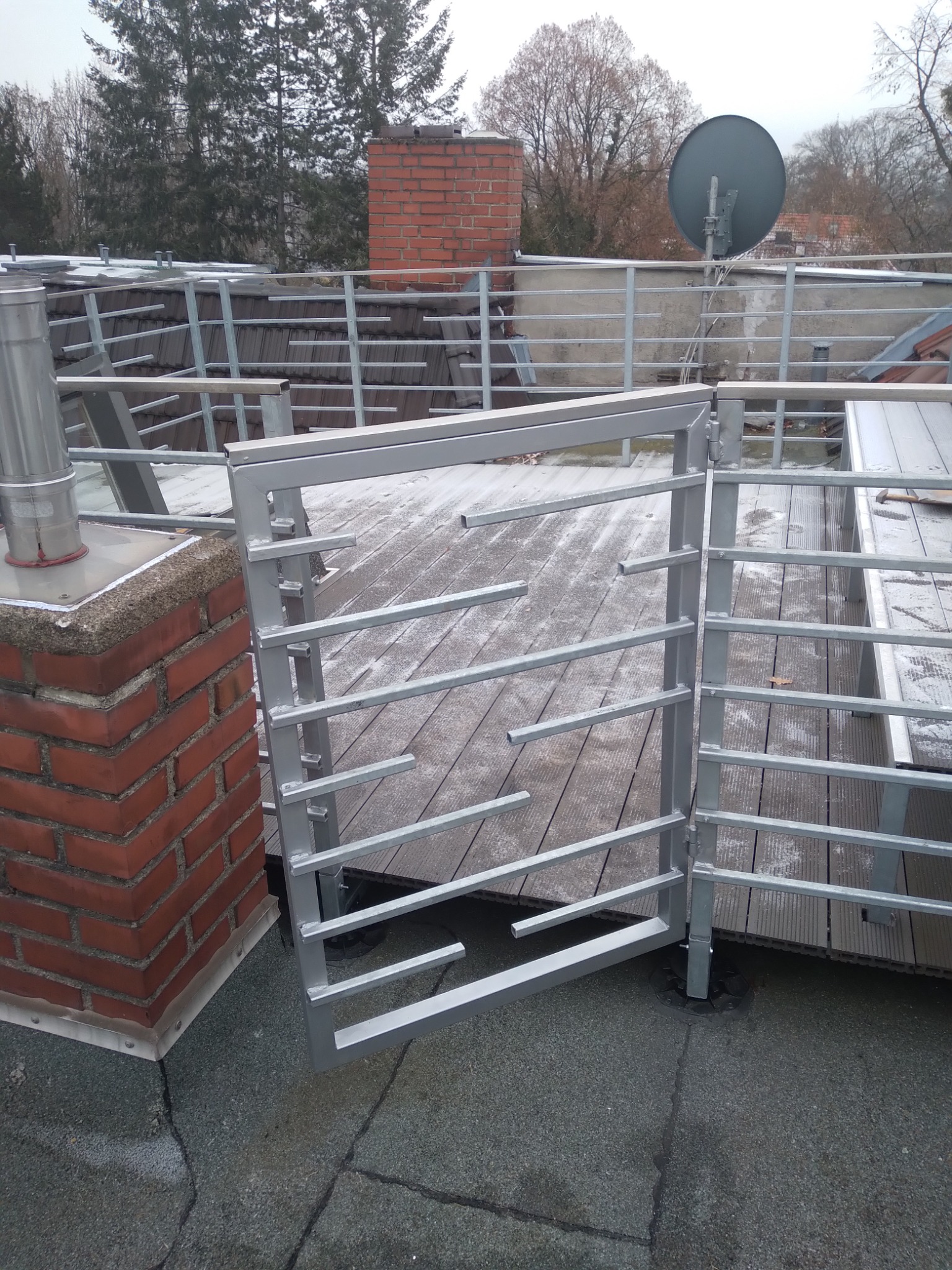 Szara metalowa balustrada na dachu z drewnianym tarasem, fragment komina z cegły i antena satelitarna w tle.