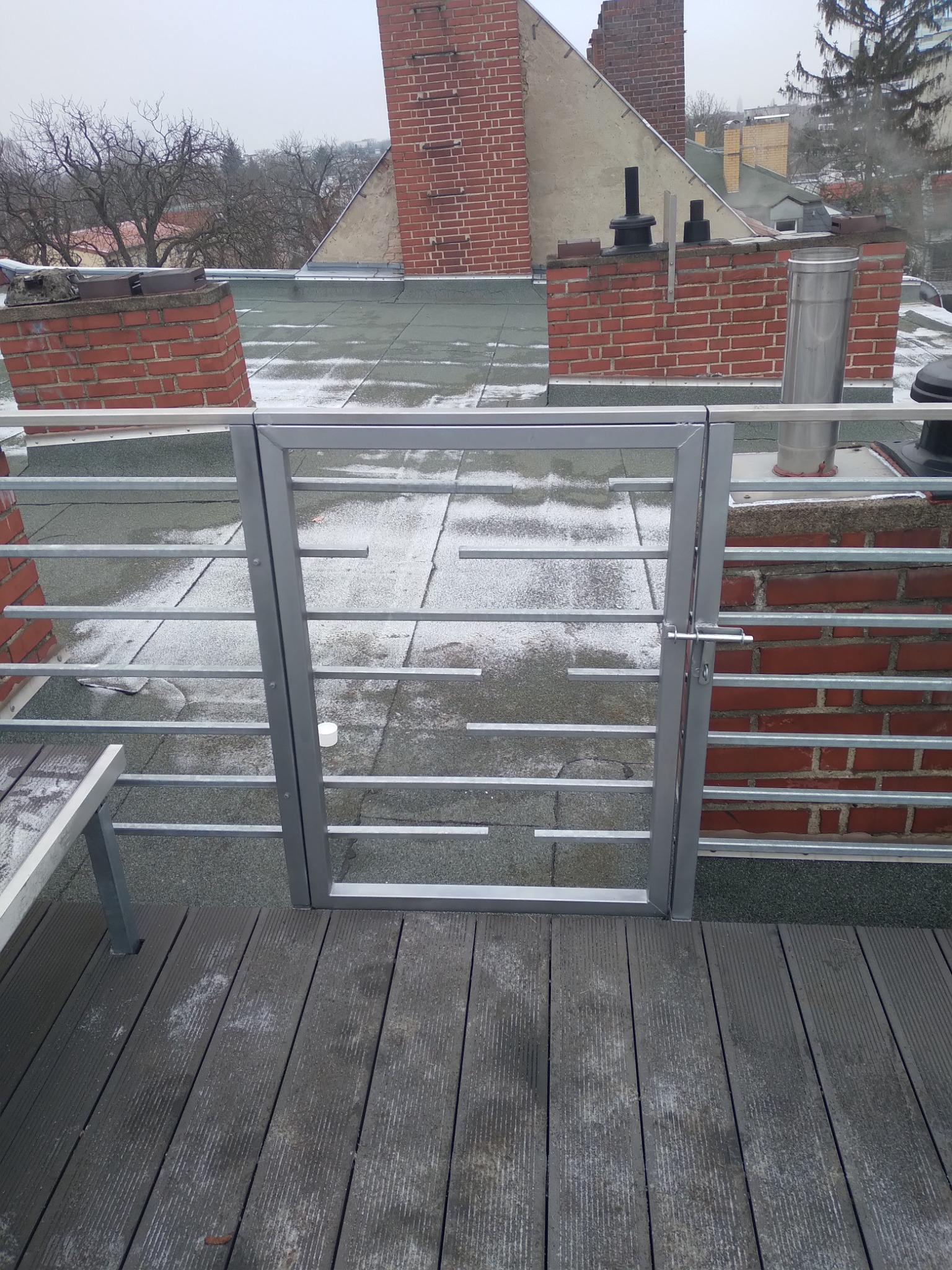 Szary metalowy płotek z furtką na dachu z drewnianym tarasem, widoczne kominy z czerwonej cegły i ośnieżone dachy budynków w tle.