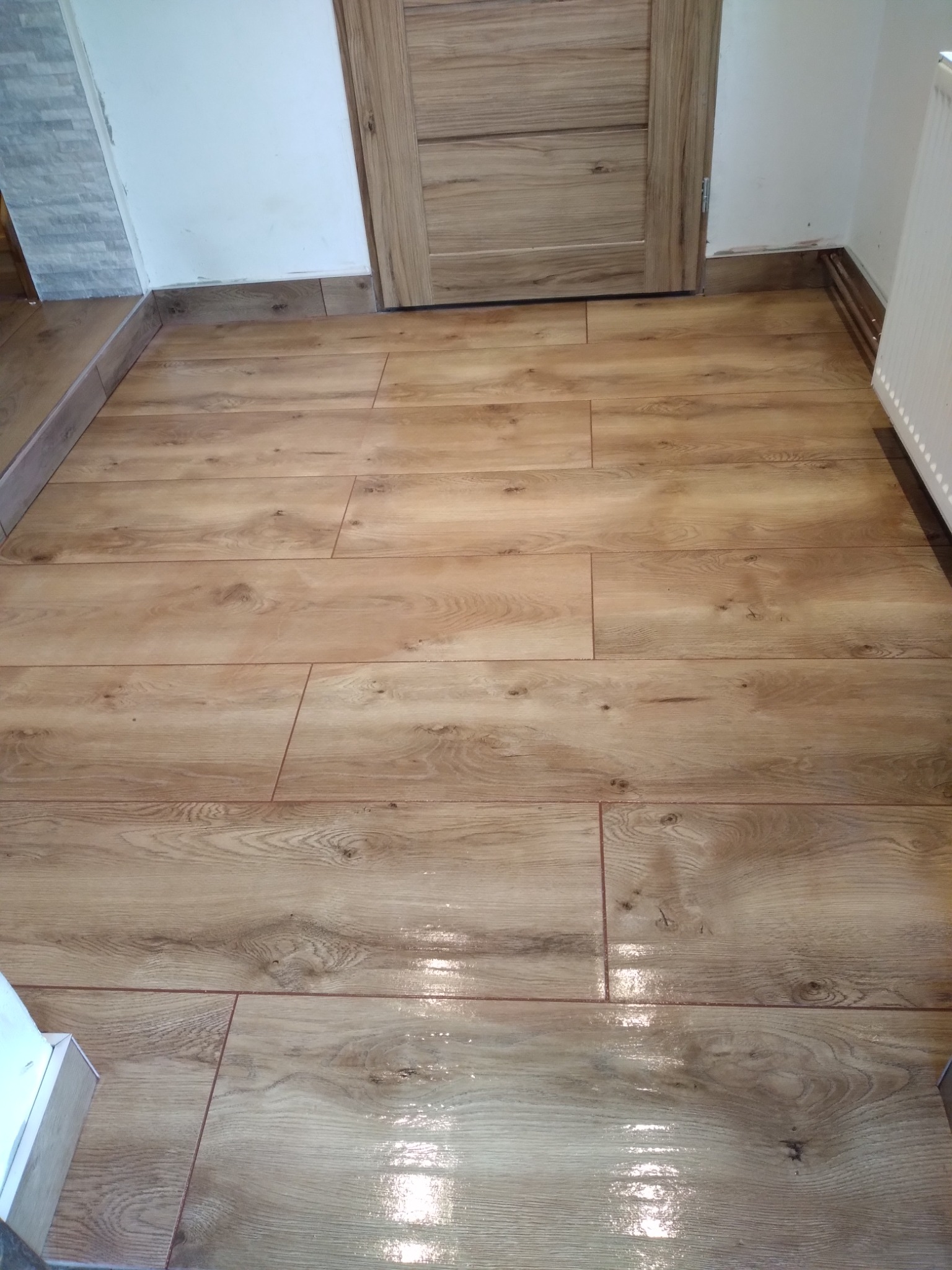 Układanie podłogi z płytek imitujących drewno w łazience, widoczna fuga w kolorze brązowym, drewniana listwa przypodłogowa i fragment grzejnika.