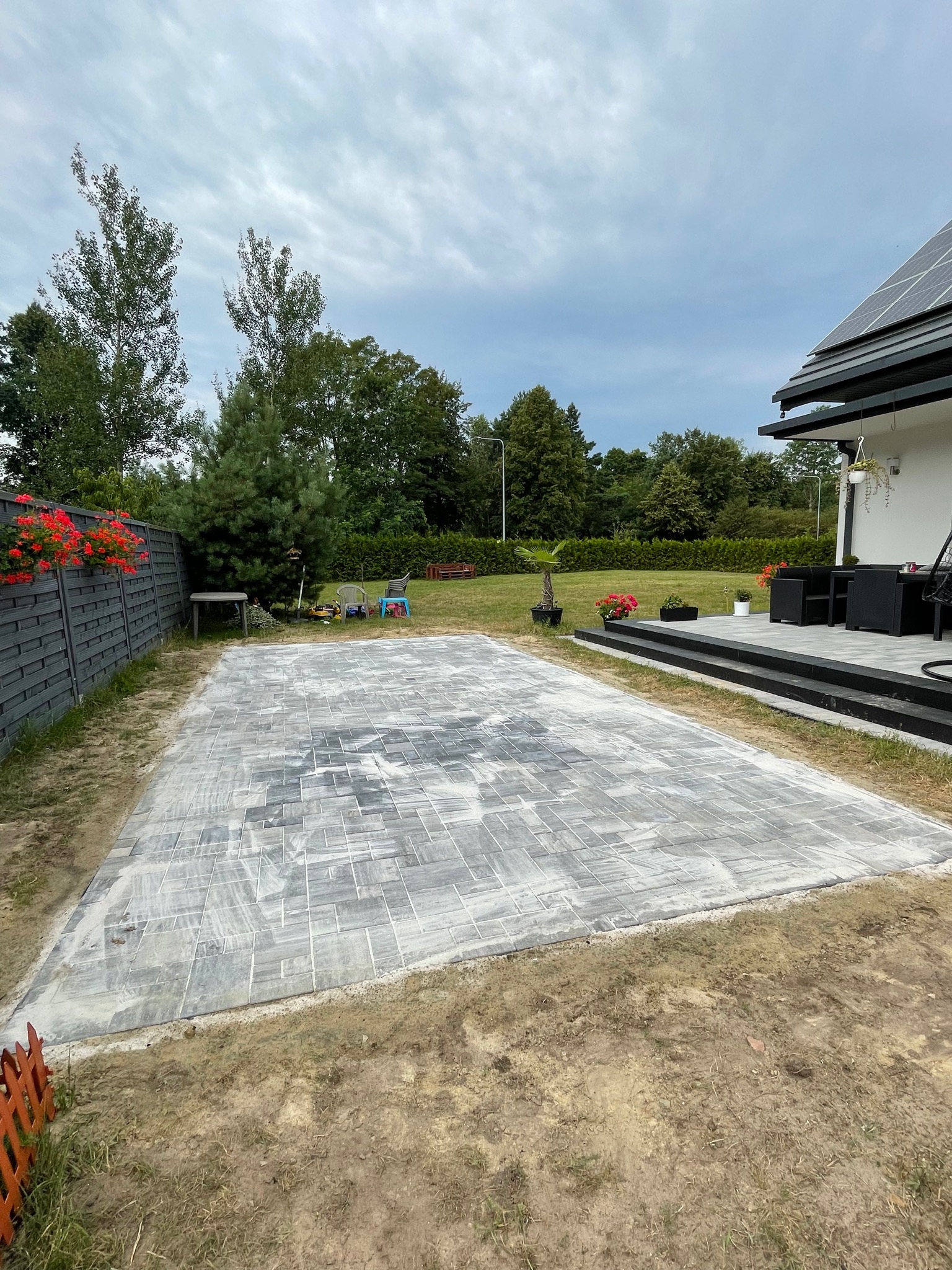 Ułożona szara kostka na przygotowanym podłożu, widok z góry na plac przydomowy, w tle dom z panelami słonecznymi i meble ogrodowe.