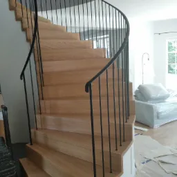 Drewniane schody spiralne z czarną, metalową balustradą, widok z dołu na górę, częściowo zakryte meble w folii ochronnej, podłoga zabezpieczona kartonem.