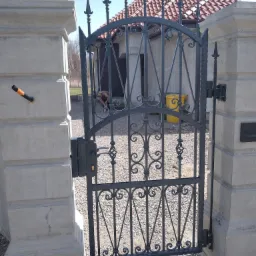 Otwarta, grafitowa brama z kutymi zdobieniami, zamontowana między dwoma betonowymi słupkami z widocznym okablowaniem elektrycznym w jednym ze słupków, na tle domu z czerwoną dachówką.