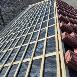 Konstrukcja drewniana dachu pokryta membraną, z przygotowanymi do ułożenia dachówkami ceramicznymi w tle, widok z góry na budynek w trakcie prac dekarskich.