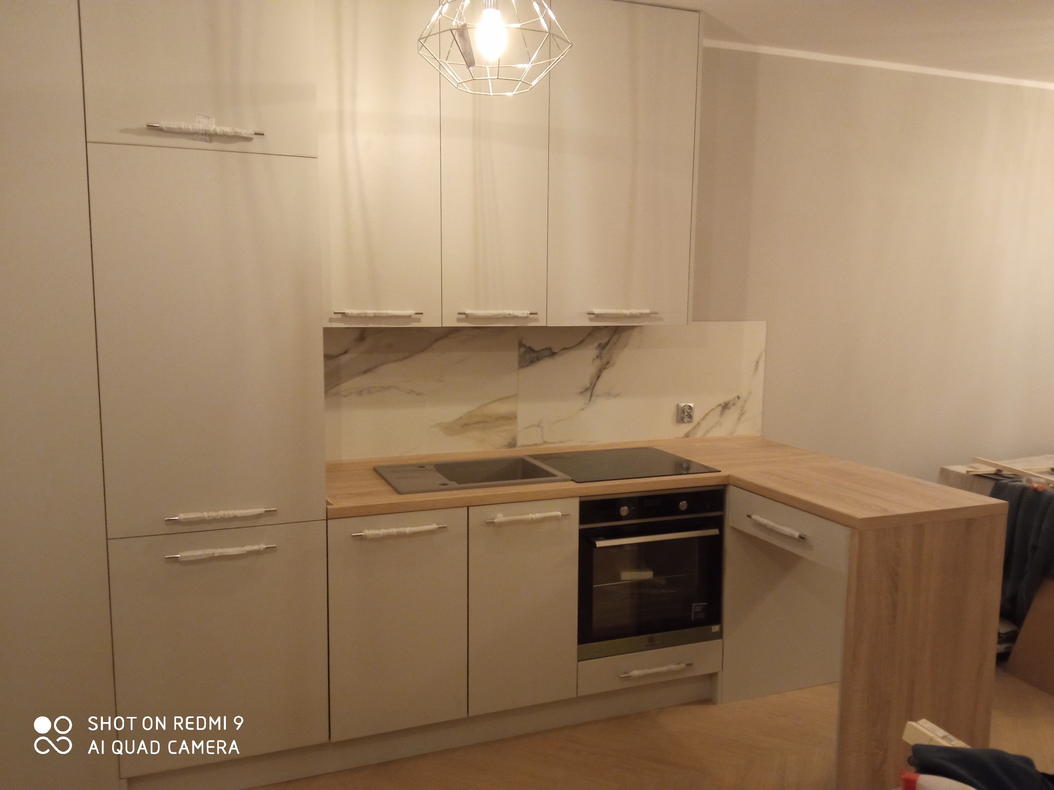 Jasna zabudowa kuchenna z drewnianym blatem, marmurowym panelem nad blatem i nowoczesną lampą wiszącą. Uchwyty szafek owinięte folią ochronną.