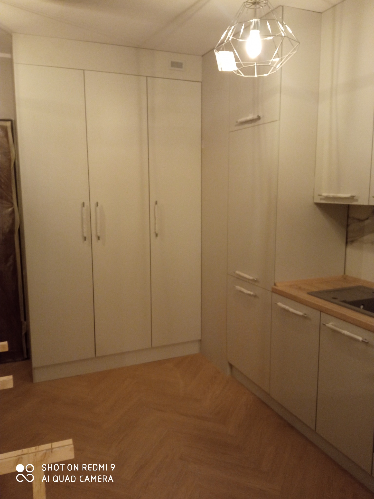 Jasna kuchnia na wymiar z drewnianym blatem i parkietem ułożonym w jodełkę, widoczny fragment niezabezpieczonej ściany w lewym rogu sugerujący trwający montaż.