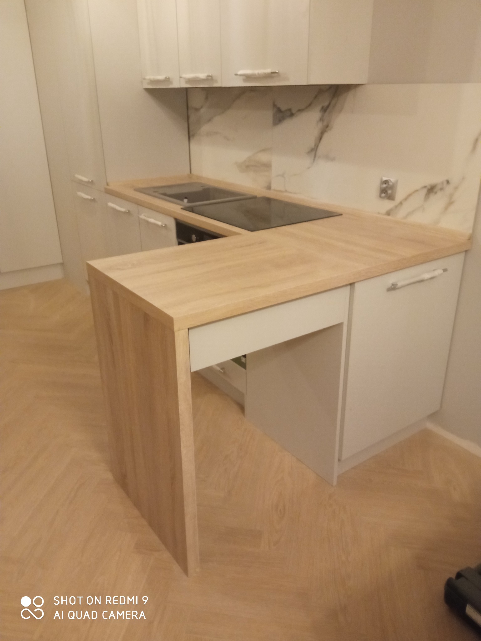 Jasna kuchnia z drewnianym blatem i marmurowym backsplashiem, widoczny półwysep kuchenny z blatem w kolorze drewna.