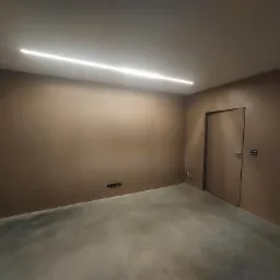 Wnętrze pokoju z brązowymi ścianami po nałożeniu gładzi, widoczna listwa LED na suficie, szary betonowy podkład, gniazdka elektryczne i minimalistyczne drzwi.