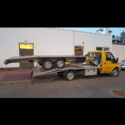 Żółty samochód dostawczy z platformą transportową przewożący przyczepę na tle białego budynku.